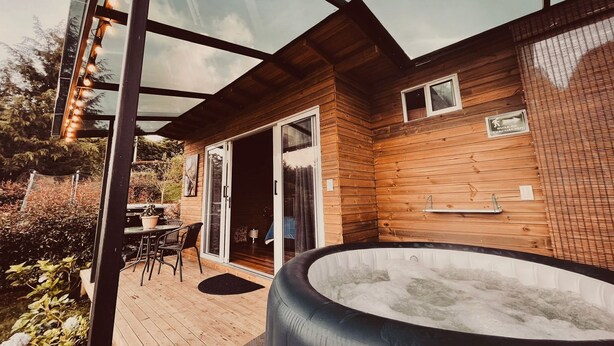A modern outdoor area with a hot tub, wooden deck, glass doors, and a seating area with chairs. - BeHappy Glamping (Medellín)