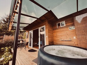 A modern outdoor area with a hot tub, wooden deck, glass doors, and a seating area with chairs. - BeHappy Glamping (Medellín)
