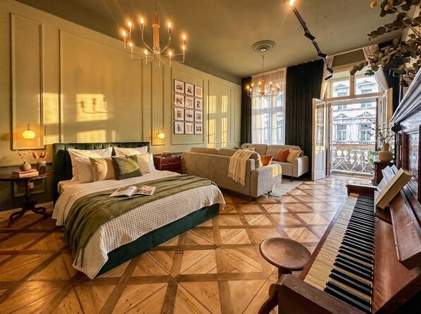 Amplia habitación de hotel con una cama grande, un sofá, un piano y un balcón con vistas a edificios.
