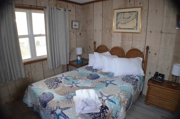 Un dormitorio con un muro revestido de madera, una cama con cubrecama floral, una mesita de noche con lámpara, y un cuadro enmarcado en la pared.