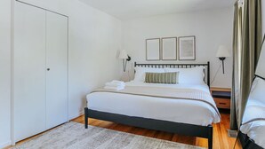 A neatly made bed with white linens and a striped pillow, a bedside table with a lamp, and a door to the left.