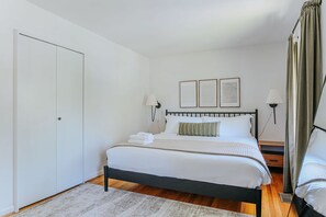 A neatly made bed with white linens and a striped pillow, a bedside table with a lamp, and a door to the left. - Westerly Haven B: Long term lease available (Charlottesville)