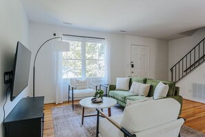 Un salon avec un canapé vert, des fauteuils blancs, une table basse et un téléviseur posé sur un meuble noir.