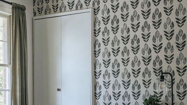 A room with a white door, a black desk with a brown chair, a potted plant, and a patterned wallpaper.
