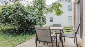 An outdoor patio with a table and chairs, surrounded by greenery and a building in the background.