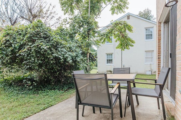 An outdoor patio with a table and chairs, surrounded by greenery and a building in the background.
