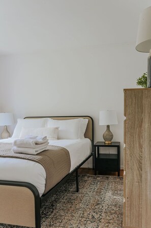 Un lit bien fait, avec du linge de lit blanc et un plaid beige, une table de chevet avec une lampe et une commode en bois.