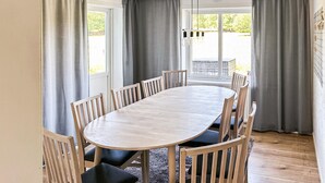 Une table ronde en bois, entourée de six chaises, avec des rideaux gris et une vue sur la verdure à l’extérieur.