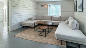 Un salon moderne avec un canapé d’angle, deux tables basses, un tapis tissé et un luminaire suspendu.