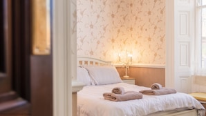 Una cama impecablemente arreglada con dos toallas enrolladas, una mesita de noche con un candelabro y un papel tapiz con motivos florales.