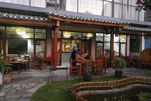 Een buiten eethoek met houten tafels en stoelen, een kleine tuin en een traditioneel gebouw met een pannendak.