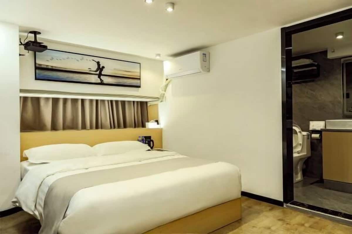 A hotel room with a bed, a painting on the wall, an air conditioning unit, and a bathroom with a toilet and sink.