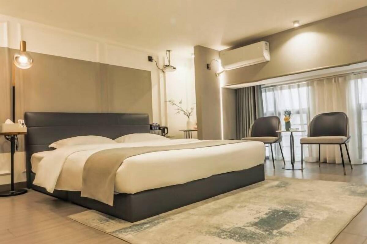 A modern hotel room with a large bed, a desk, two chairs, a small table, and a large window with curtains.