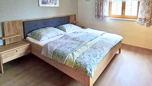 Ein Einzelbett mit hölzernem Gestell und Kopfteil, ein Nachttisch und ein Fenster mit Vorhängen.