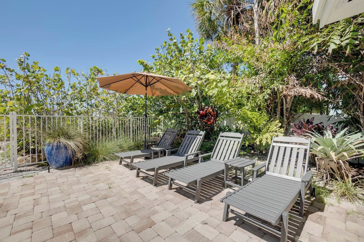 A patio with lounge chairs and a large umbrella, surrounded by lush greenery and a white fence.
