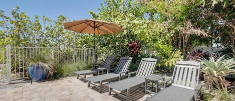 A patio with lounge chairs and a large umbrella, surrounded by lush greenery and a white fence.