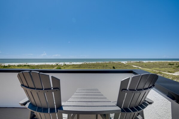 A balcony with two Adirondack chairs and a table overlooking a beach and ocean.