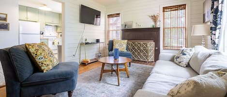 A living room with a sofa, armchair, and a coffee table. There is a TV mounted on the wall and a window with curtains.