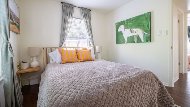 A bedroom with a bed, a nightstand, a lamp, a window with curtains, and a painting on the wall. - Vintage Rosedale Bungalow in Rosedale (Austin)