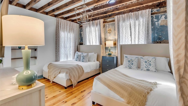 Une chambre avec deux lits, un plancher en bois, un lustre et une grande fenêtre avec des rideaux.