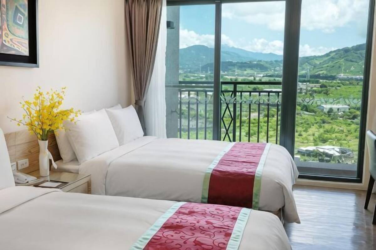 Ein Hotelzimmer mit zwei Betten, einem Nachttisch, einer Vase mit gelben Blumen und einem großen Fenster mit Blick auf grüne Hügel.