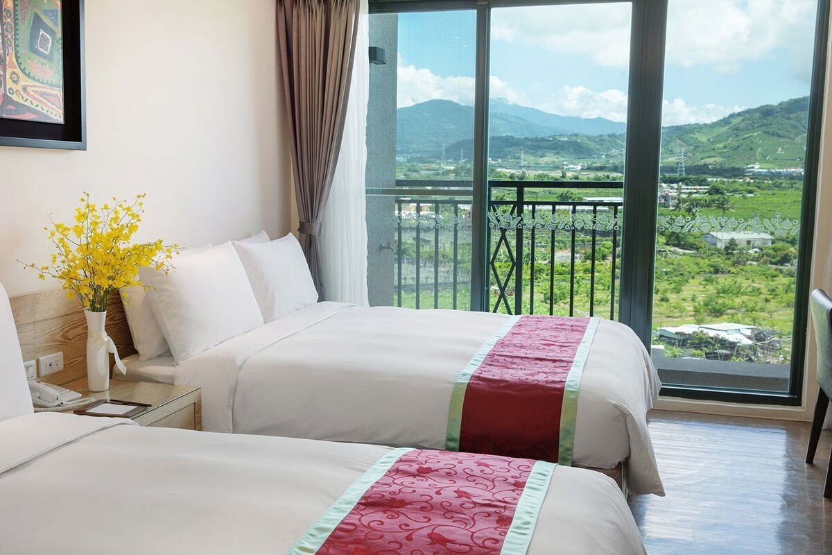 A hotel room with two beds, a bedside table with a phone and a vase of yellow flowers, and a large window offering a view of green hills and a distant cityscape.