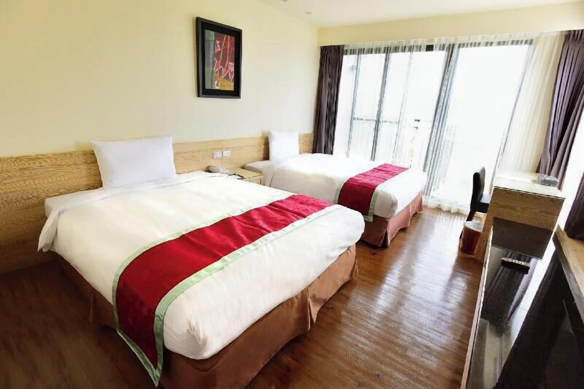 A hotel room with two beds, a wooden floor, a window with curtains, and a painting on the wall.