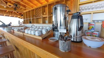Una barra de desayuno con máquinas de café, una bandeja de tazas blancas, un tazón de azúcar y bebidas enlatadas.