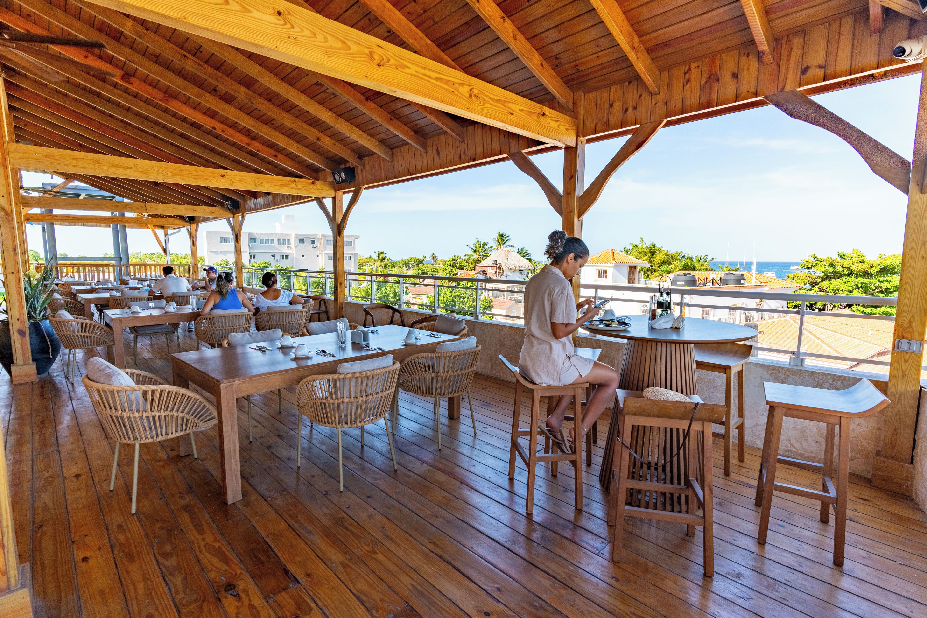 An outdoor wooden deck with tables and chairs, overlooking a coastal area with buildings and palm trees.