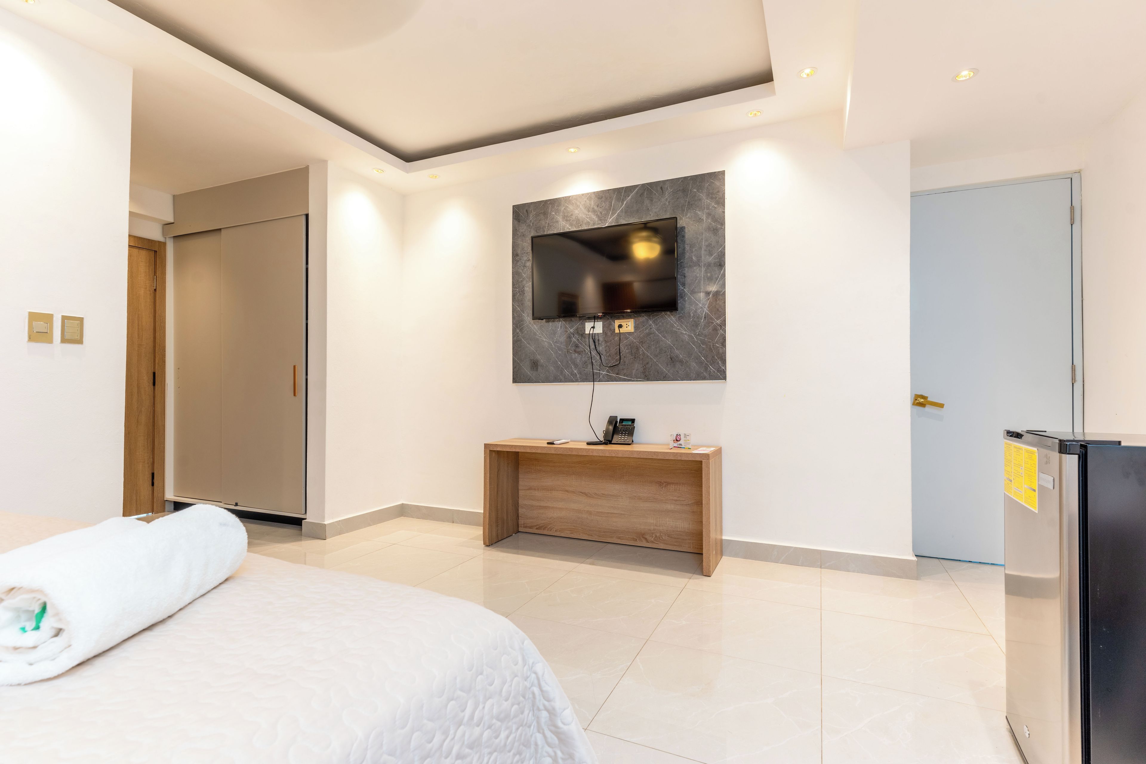 A modern hotel room with a bed, a wooden console table, a flat-screen TV mounted on a marble wall, a built-in refrigerator, and a door with a handle.