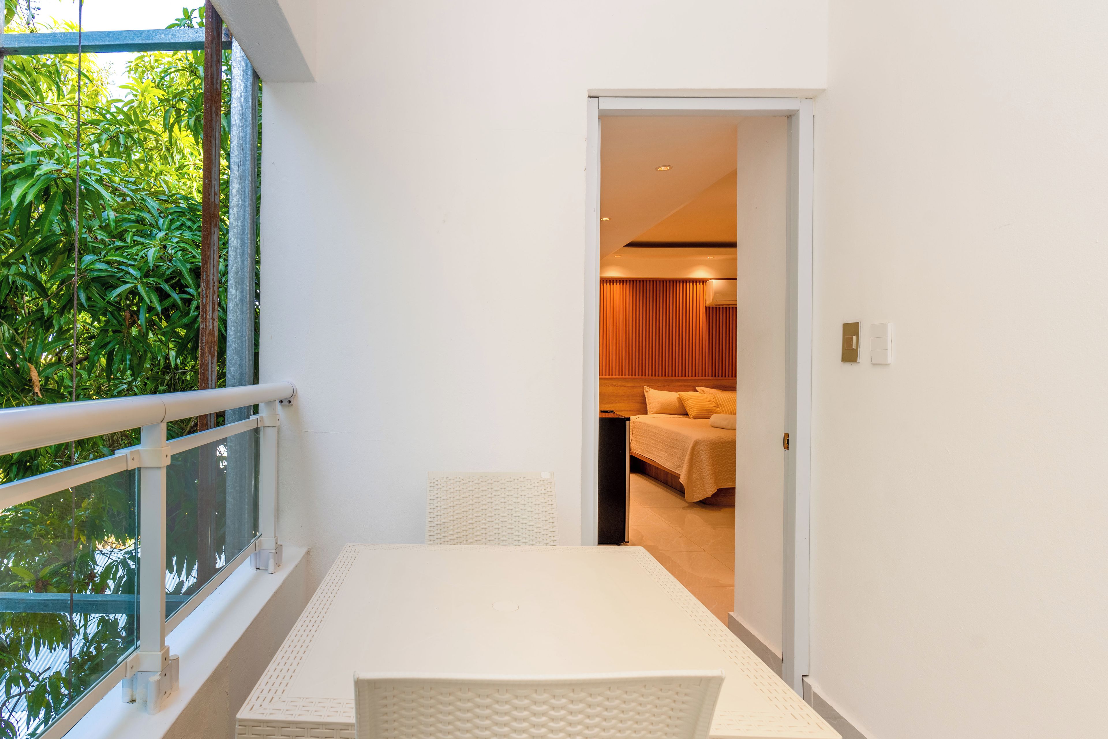 Un balcon avec une table et des chaises blanches, une balustrade en verre et une vue sur la verdure.