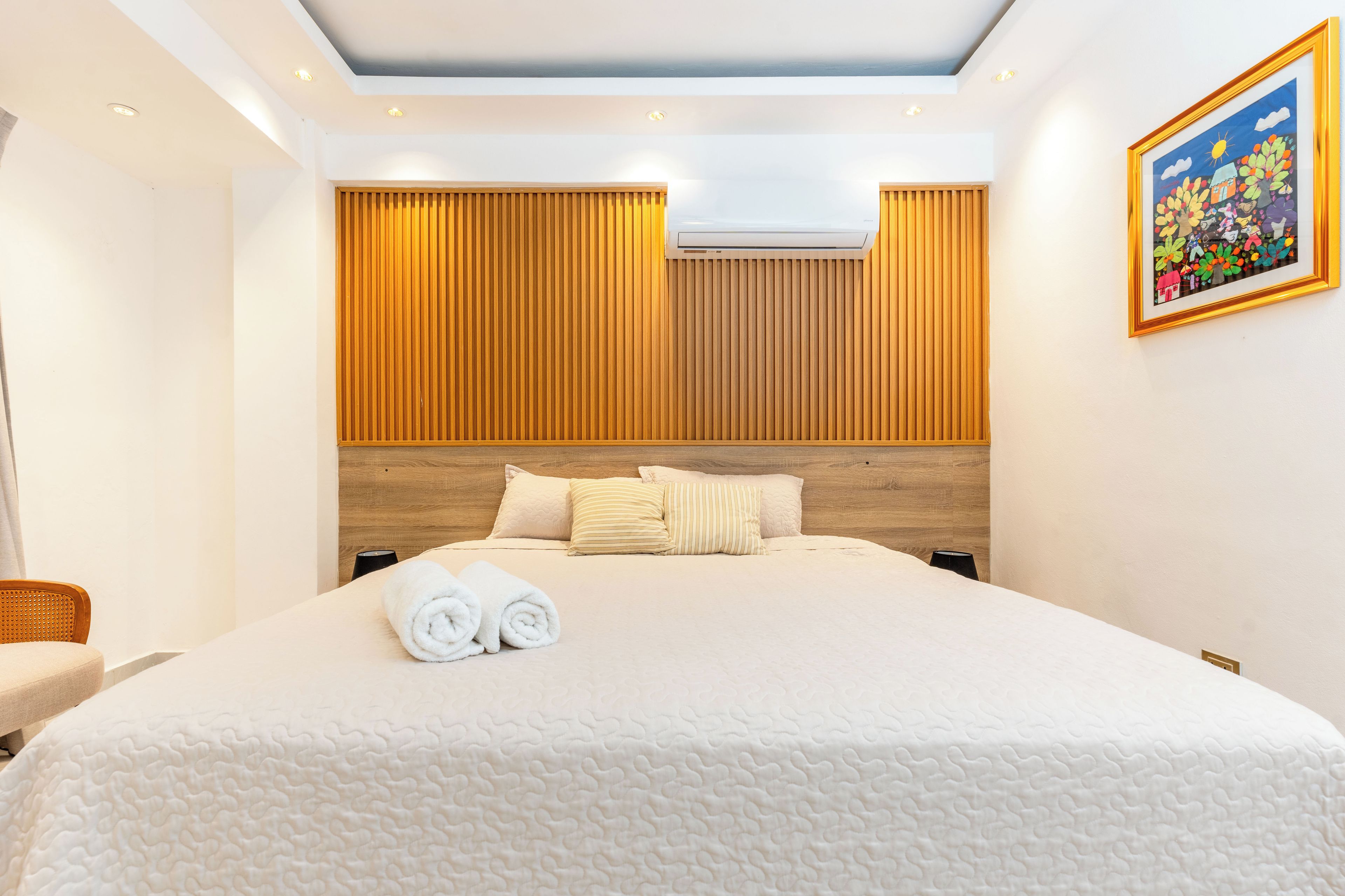 A hotel room with a large bed, a wooden headboard, an air conditioning unit, and a painting on the wall.