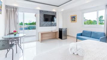 A hotel room with a bed, a desk, a chair, a TV, a small fridge, and a window with a view of greenery.