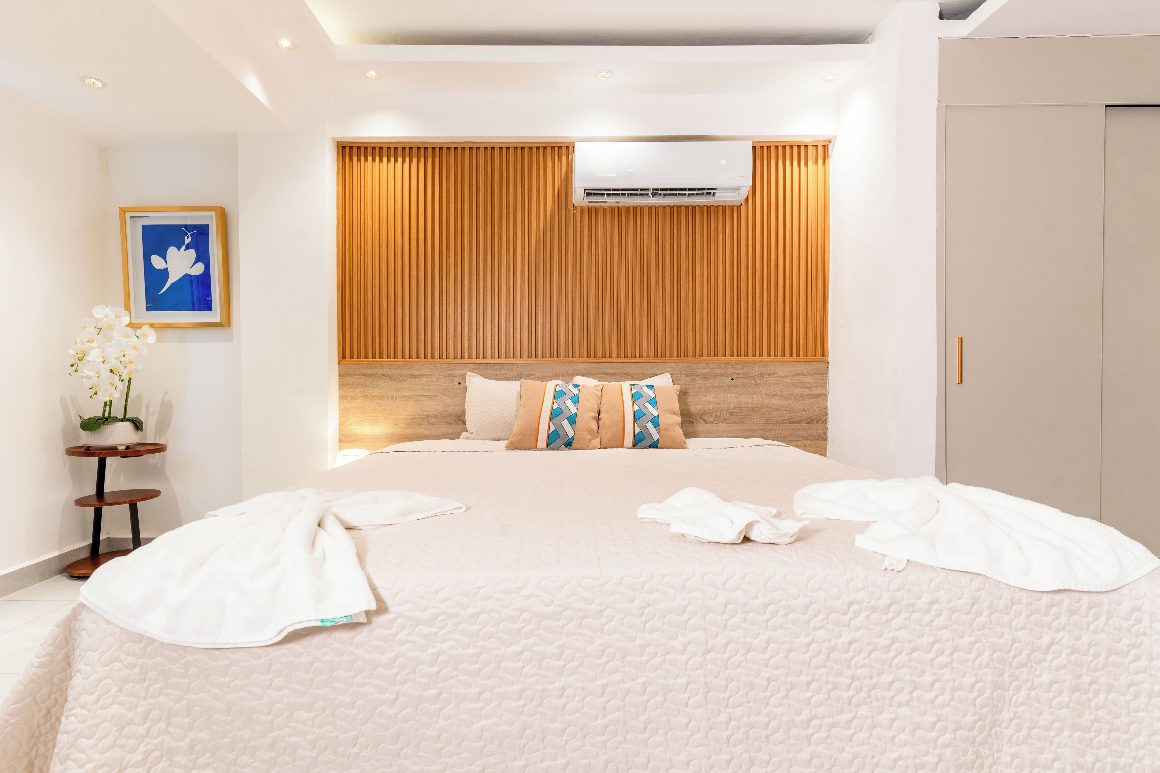 A hotel room with a large bed, a wooden headboard, an air conditioning unit, a framed picture on the wall, and a small table with a vase of flowers.