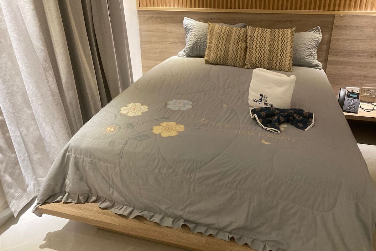 A neatly made bed with a grey bedspread featuring floral embroidery, a white towel, and a folded garment on the side.