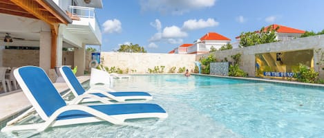 A clear swimming pool with lounge chairs, a stone wall, and a building with a red roof in the background.
