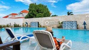 Un área de piscina con sillas de descanso, un cartel que dice 'CASUAL On the Beach BAYAHIBE', y un edificio con techo rojo al fondo.
