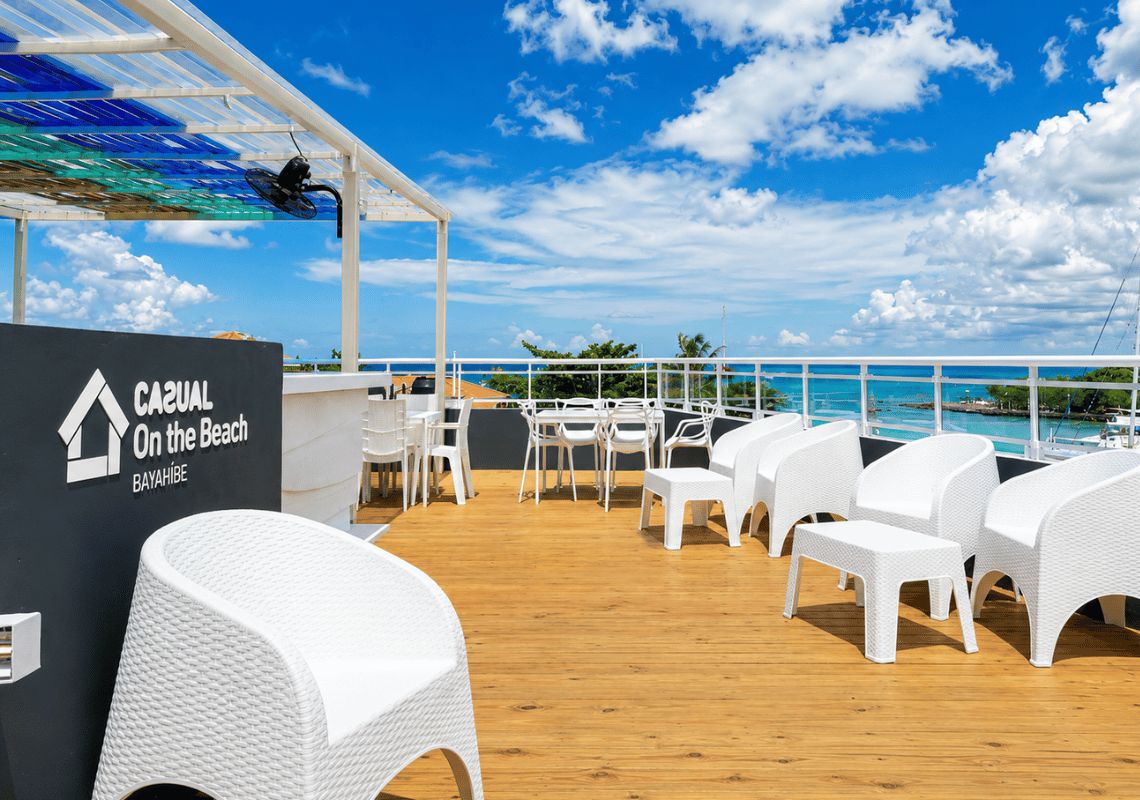 Eine Terrasse mit weißen Korbmöbeln, einem Schild mit der Aufschrift 'CAJUAL On the Beach BAYAHIBE' und Blick auf den Ozean.
