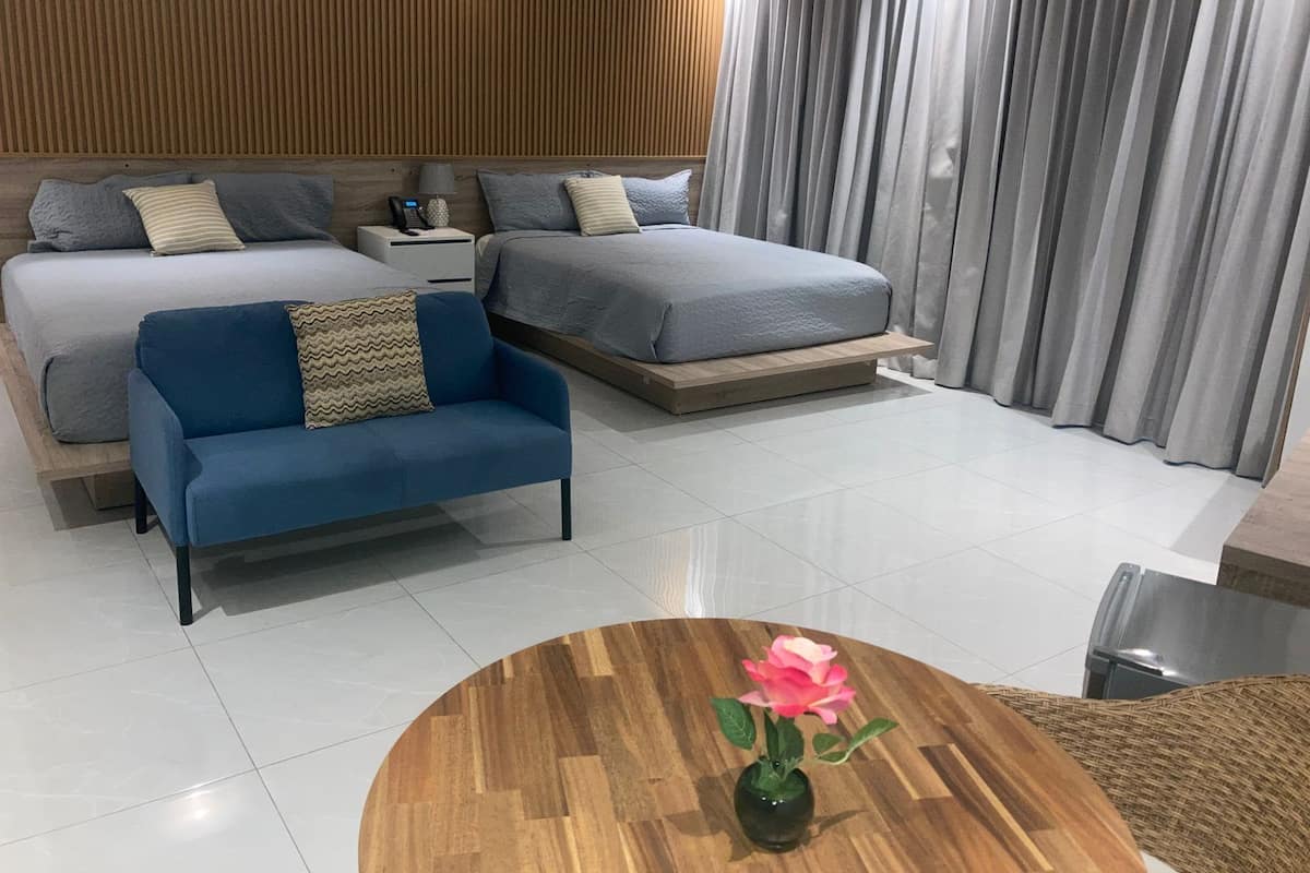 A modern hotel room with two beds, a sofa, a wooden coffee table with a vase, and a view of the city through large windows.