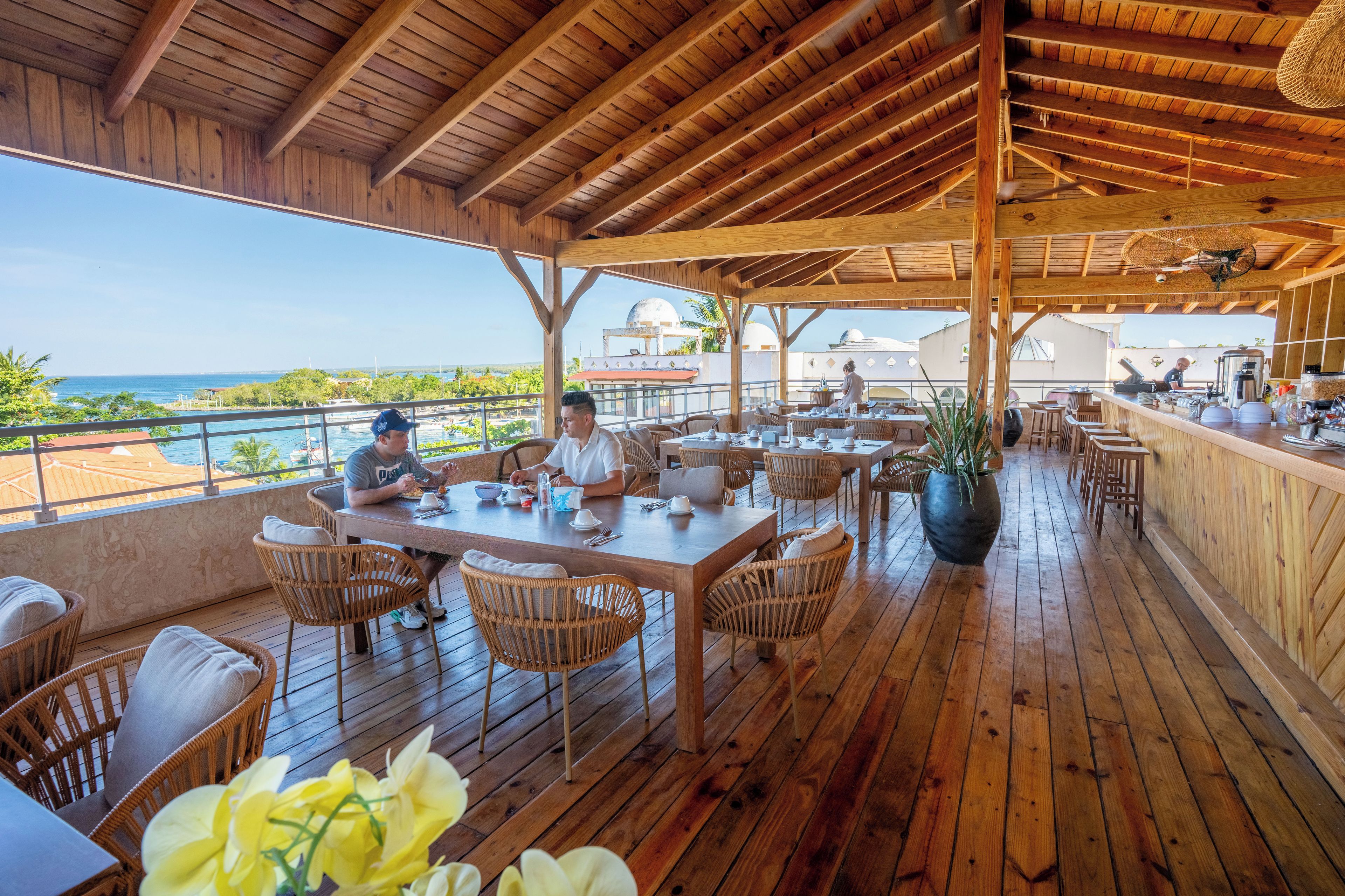 Un espace de restauration en plein air avec des tables et des chaises en bois, un bar et une vue sur l’océan.