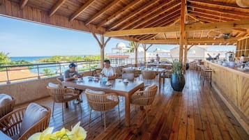 Un espace de restauration en plein air avec des tables et des chaises en bois, un bar et une vue sur l’océan.