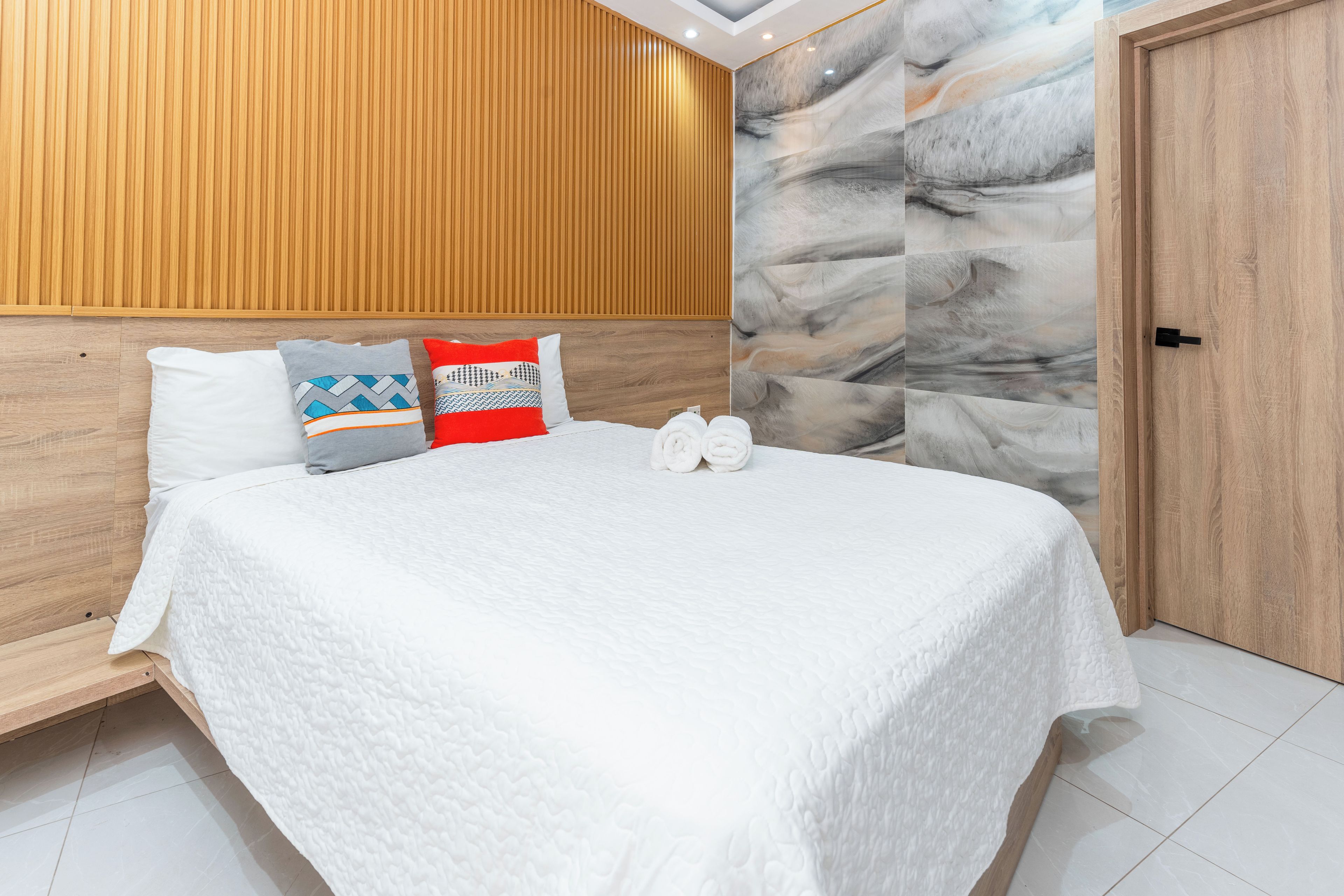Ein ordentlich bezogenes Bett mit weißer Bettwäsche und mehreren Kissen, ein Holz-Kopfstück und eine Wand mit Marmoroptik.
