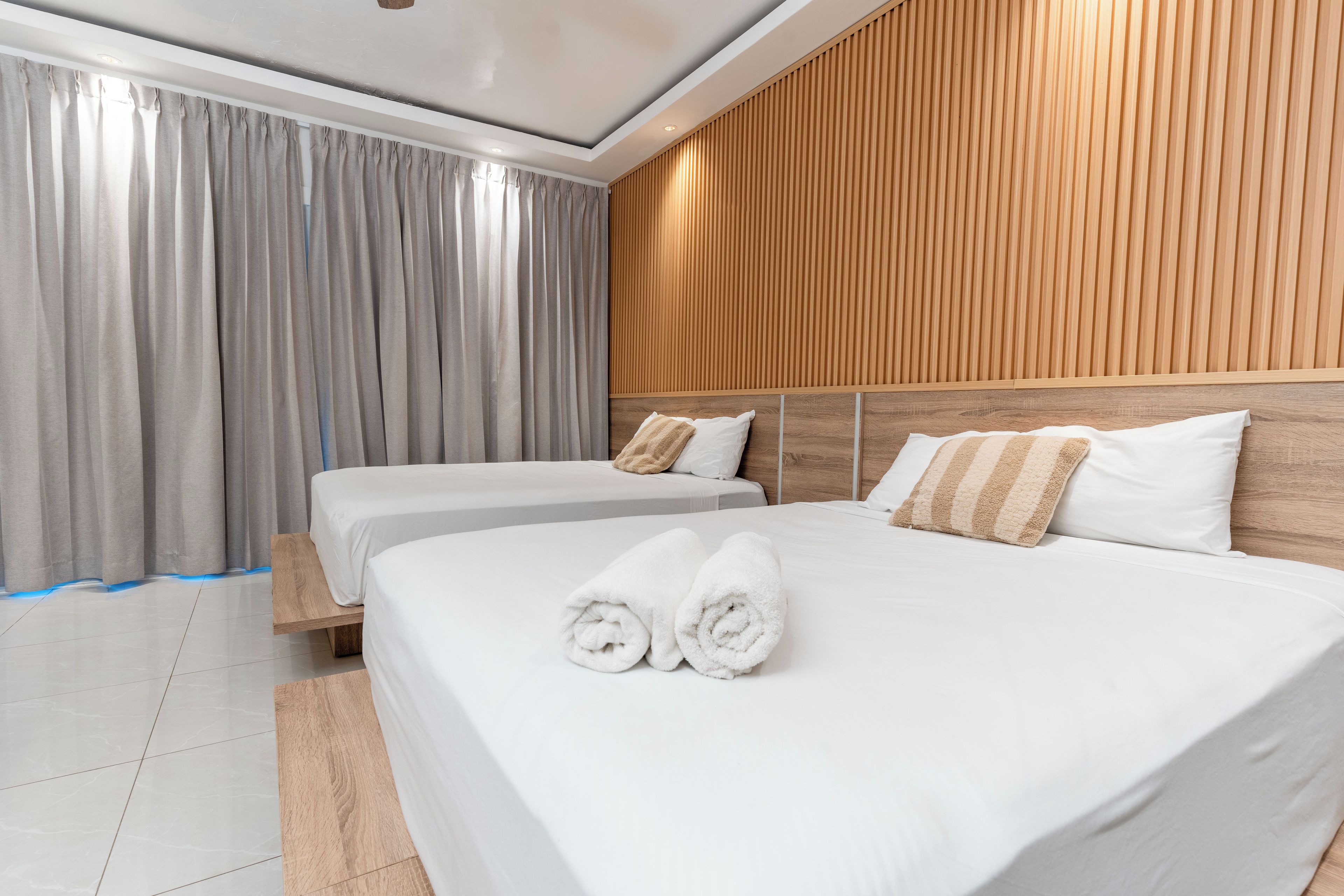 Une chambre d’hôtel avec deux lits, une tête de lit en bois, des rideaux gris et un sol carrelé.