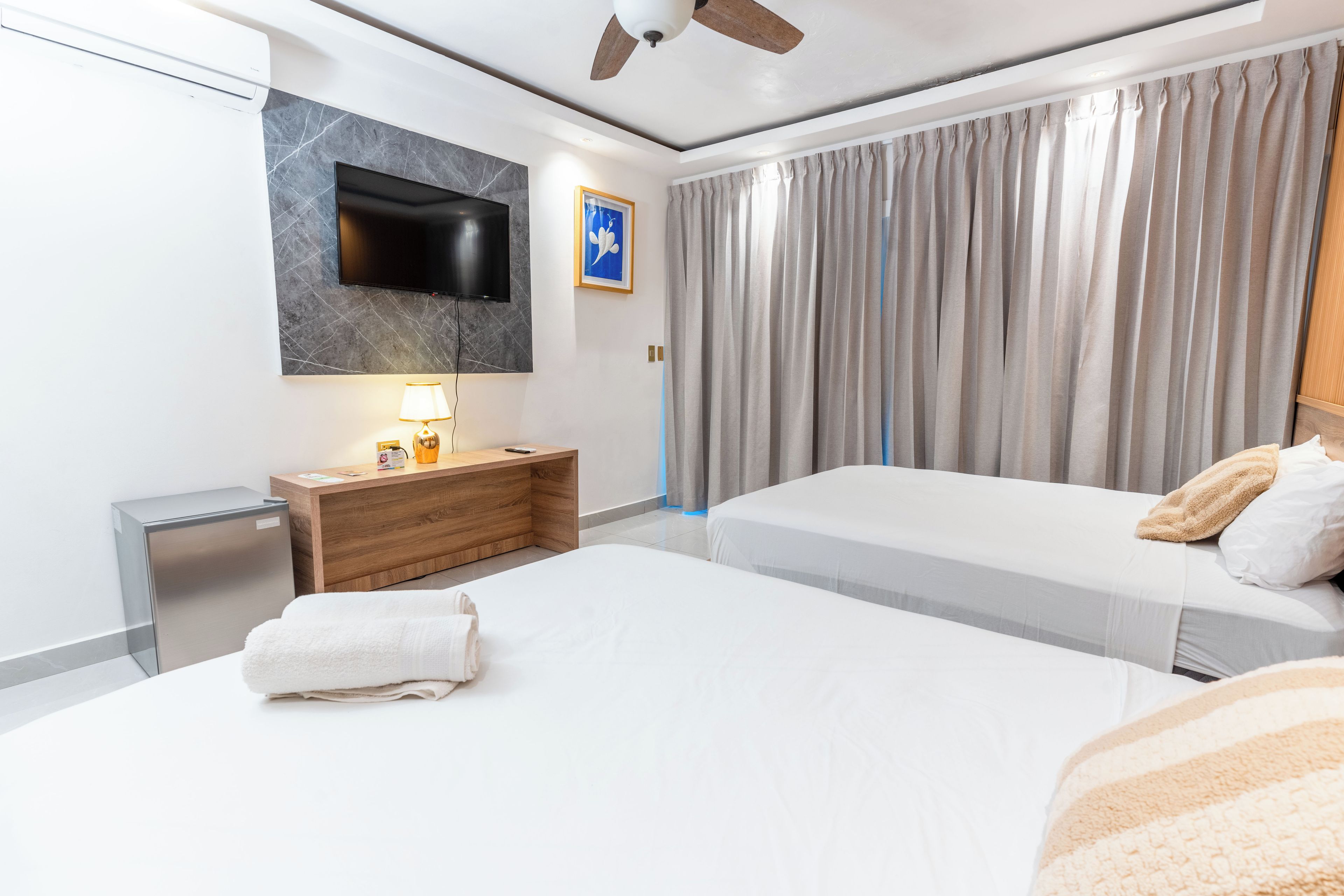 A hotel room with two beds, a TV mounted on the wall, a wooden nightstand, a desk, and a large window with curtains.