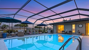 A swimming pool with lounge chairs and umbrellas, surrounded by a covered area with a view of the sunset.