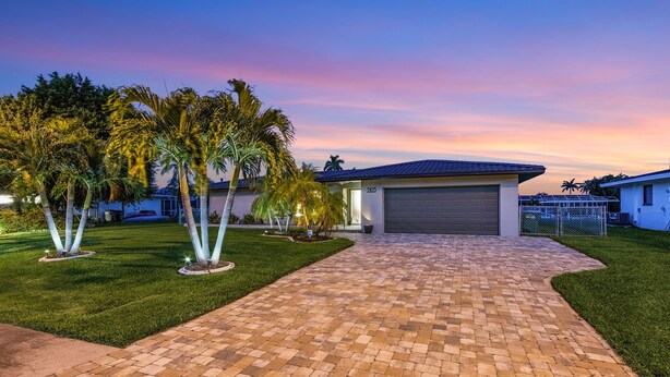 A residential property with a paved driveway, a garage, and a well-maintained lawn with palm trees.