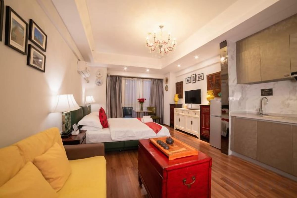 Habitación de hotel con cama, sofá, cómoda roja, zona de cocina y candelabro.