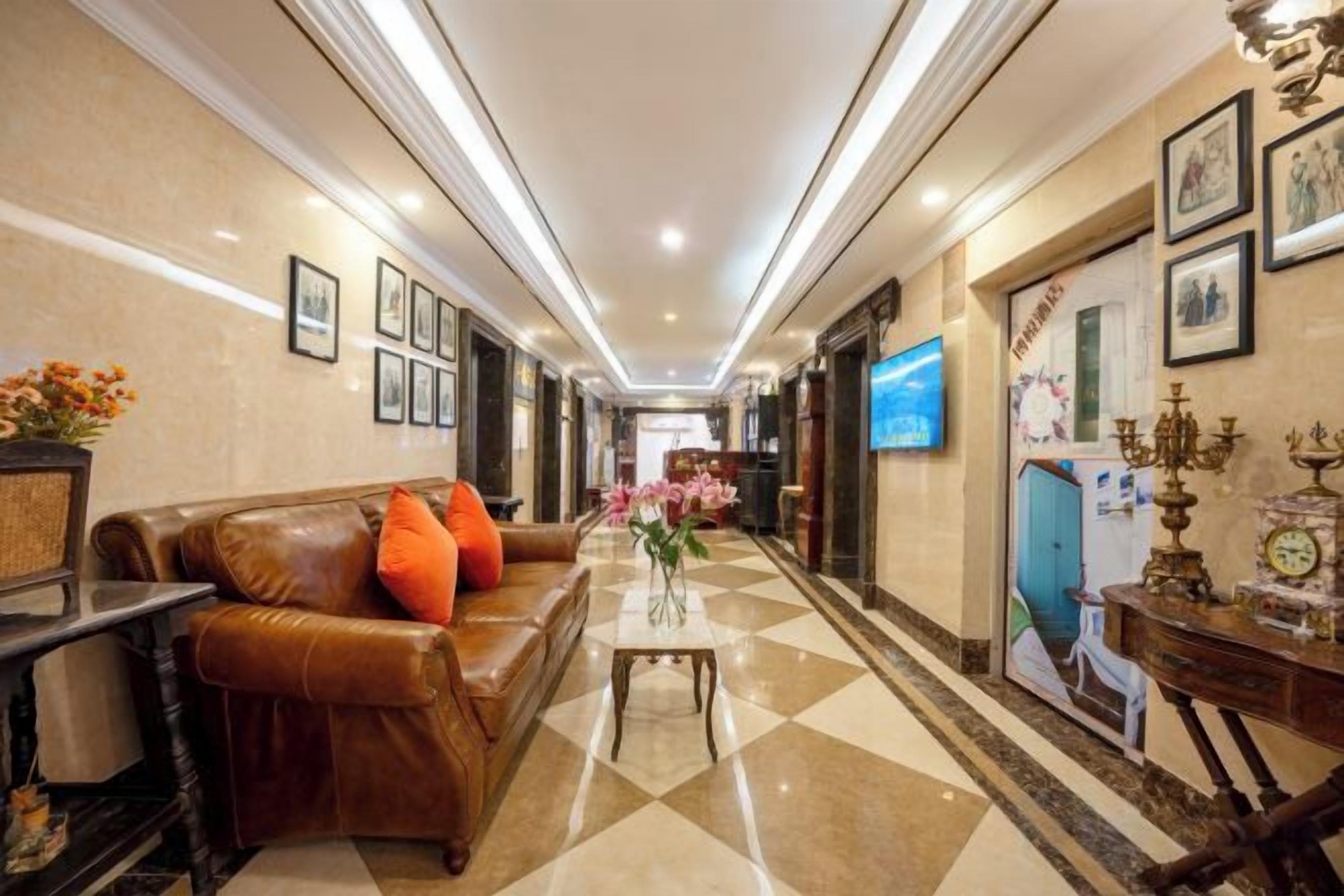 A hotel lobby with a checkered floor, a leather sofa with orange cushions, a vase with flowers, and a television on the wall.