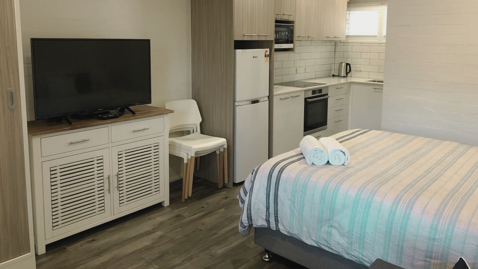 A compact room with a bed, a TV on a wooden cabinet, a small kitchenette, and a chair.