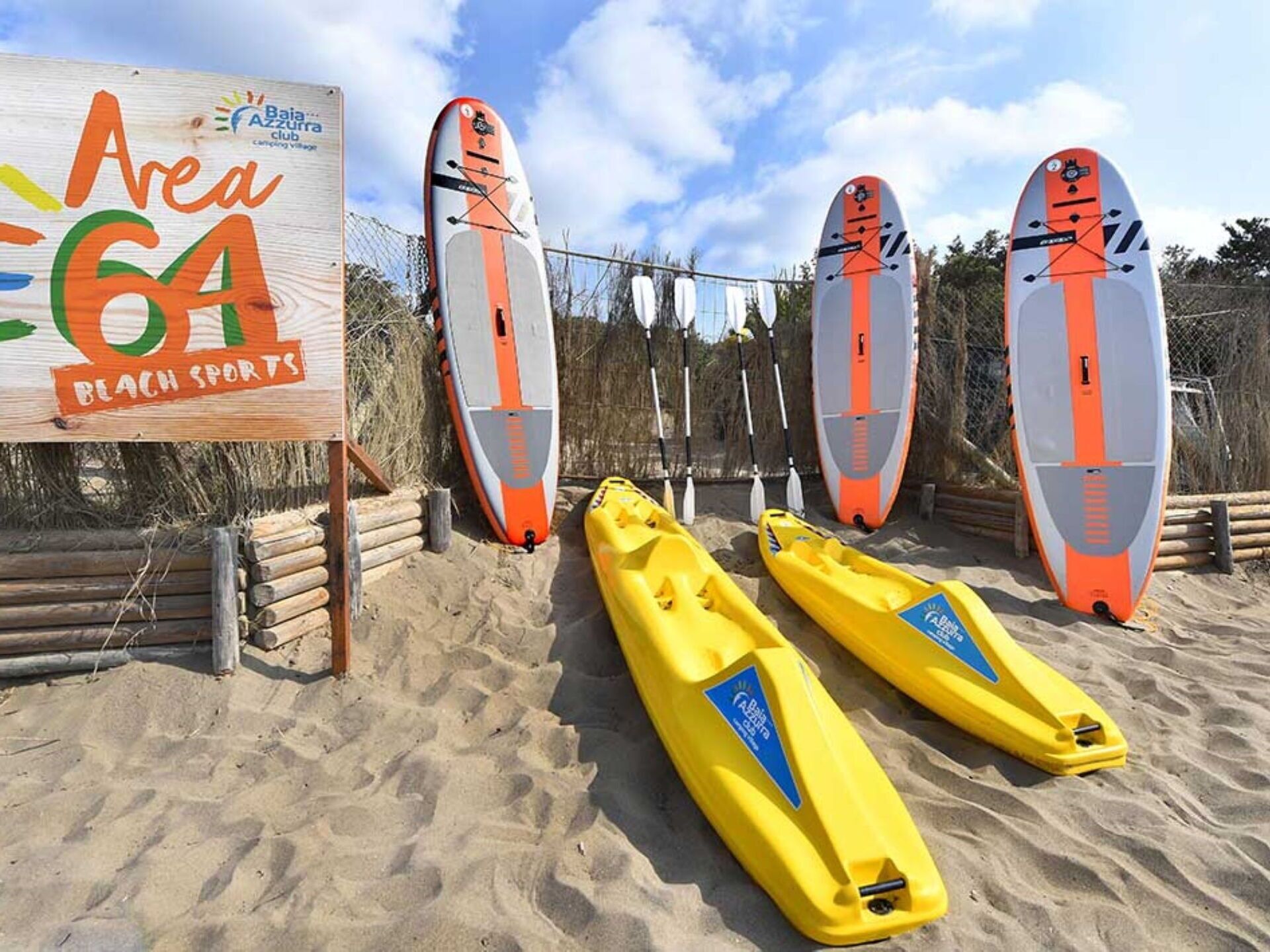 Ein Strand mit vier Stand-Up-Paddles und zwei Kajaks, einem Schild für 'Area 64 Beach Sports' und einem Holzzaun.